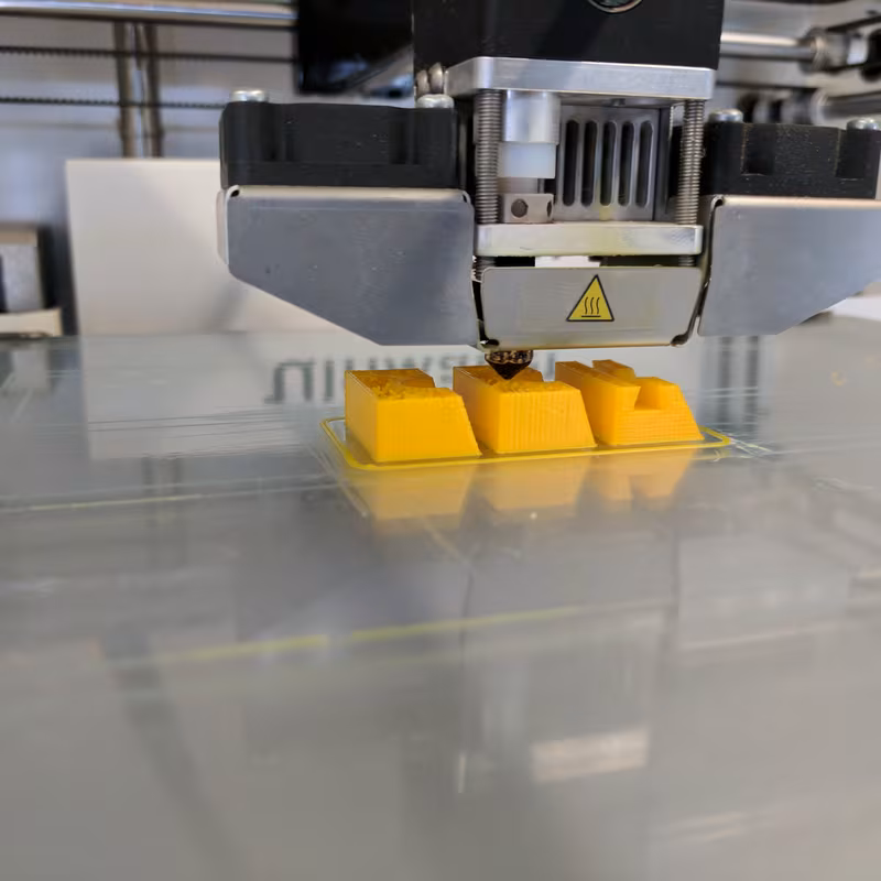 An Ultimaker print head laying down the first layers of three small yellow parts on the glass bed