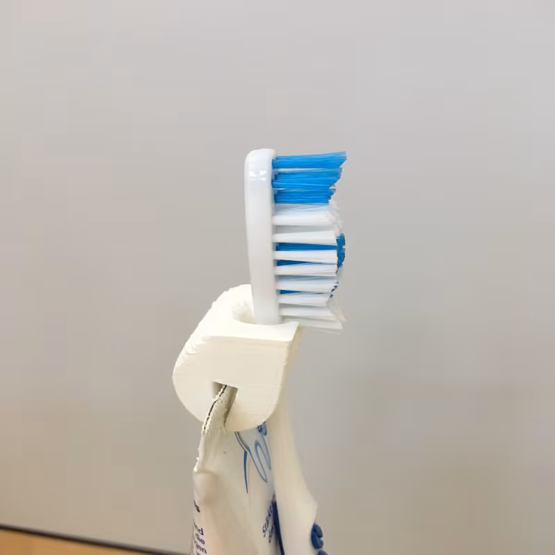 The white prototype in use, clipped to the seam of a toothpaste tube and holding a white toothbrush upright