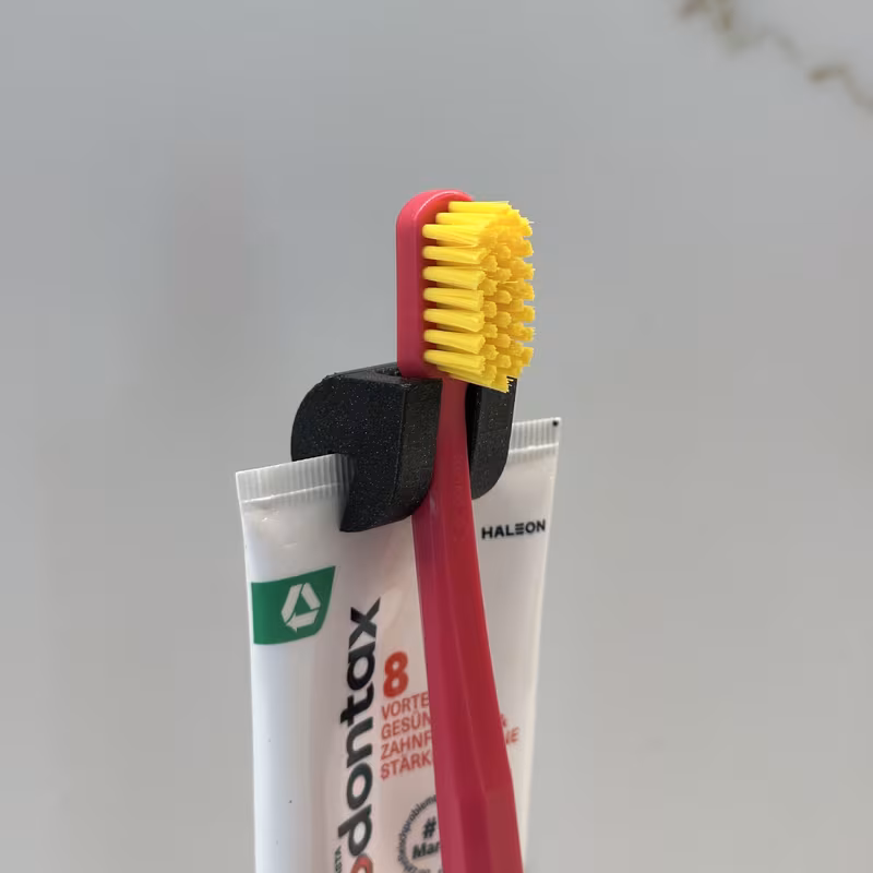 Close-up from the front of the black holder gripping the tube seam with the toothbrush neck seated in the channel