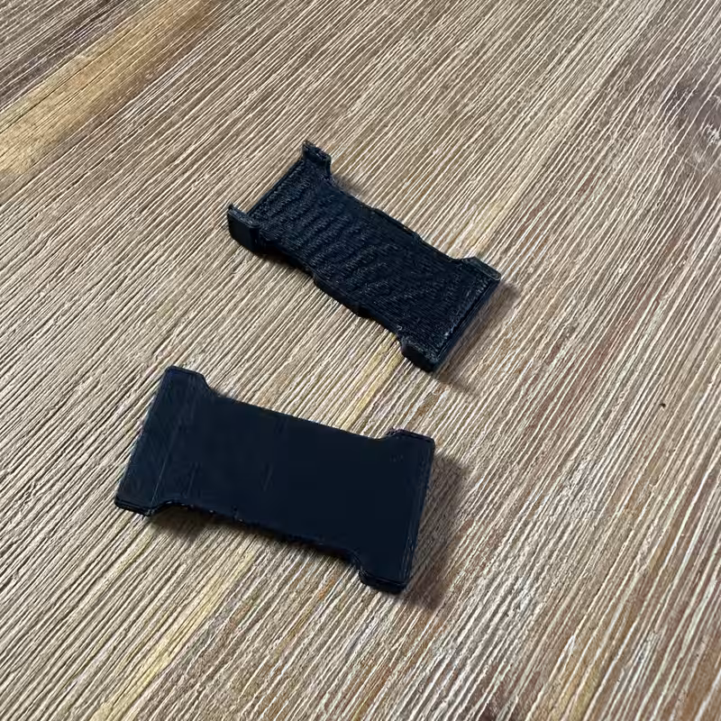 Two black 3D-printed versions of the part on a wooden surface, a thin first attempt and a thicker revision