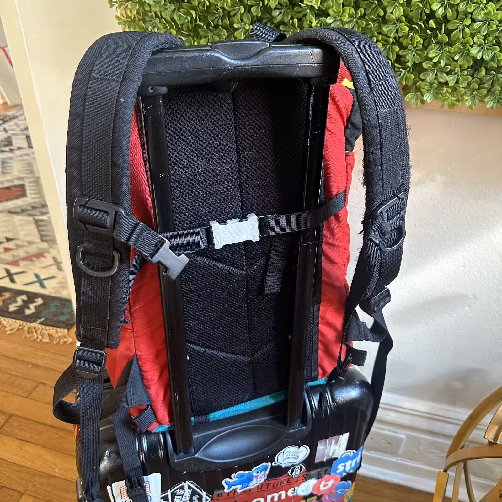 The red backpack mounted on the back of a rolling suitcase by its shoulder straps, with the white part joining a strap across the middle