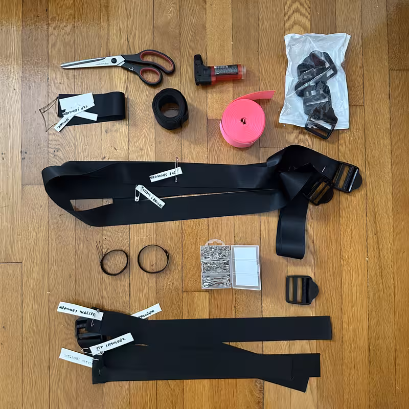 Materials laid out on a wooden floor: black seatbelt webbing, a coil of hot pink webbing, buckles in a bag, scissors, a seam ripper, safety pins, and strap pieces labelled with masking tape