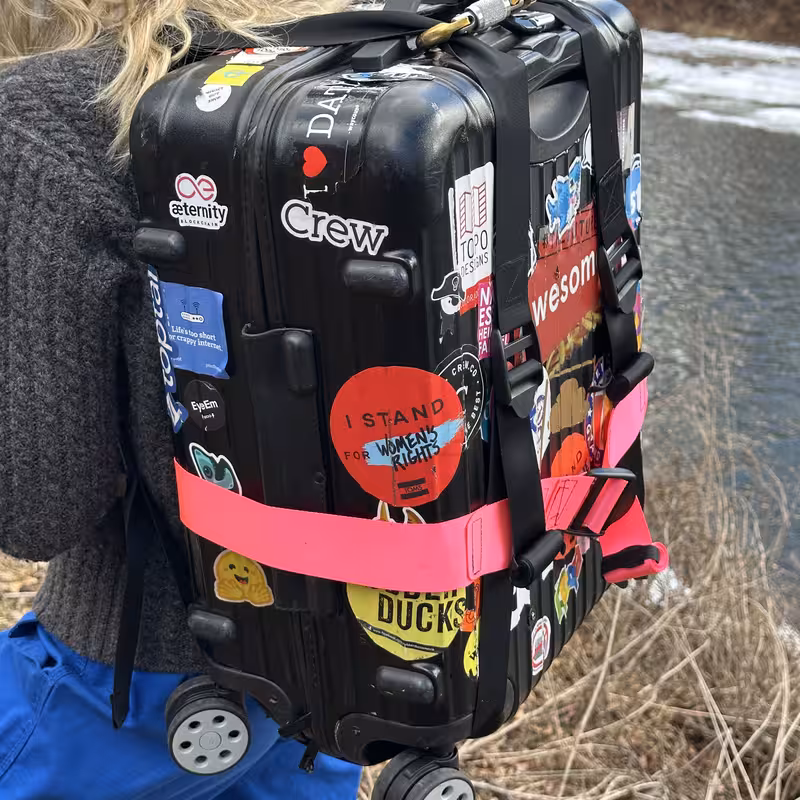 Side view of the sticker-covered case riding on the back, with the pink strap wrapped around it and the black shoulder strap running over the shoulder